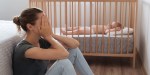 młoda matka siedzi przy łóżeczku swojego dziecka skrywając twarz w dłoniach i przeżywa depresję poporodową